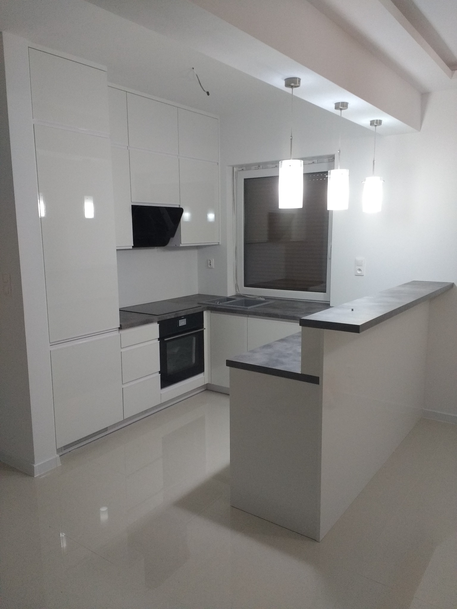 Nowoczesna, minimalistyczna kuchnia z białymi, lakierowanymi frontami, szarym blatem i wyspą kuchenną, oświetlona trzema wiszącymi lampami z mlecznego szkła.