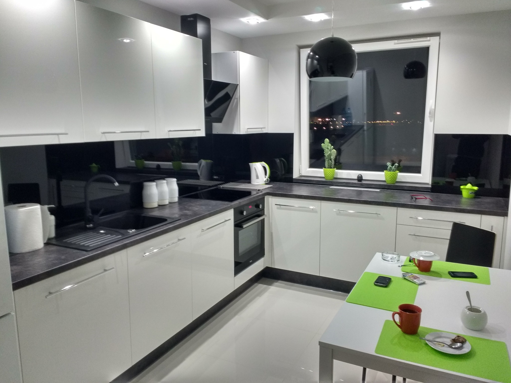 Nowoczesna, jasna kuchnia z białymi szafkami i czarnym blatem, widok na nocne miasto przez okno, stół z resztkami po posiłku.