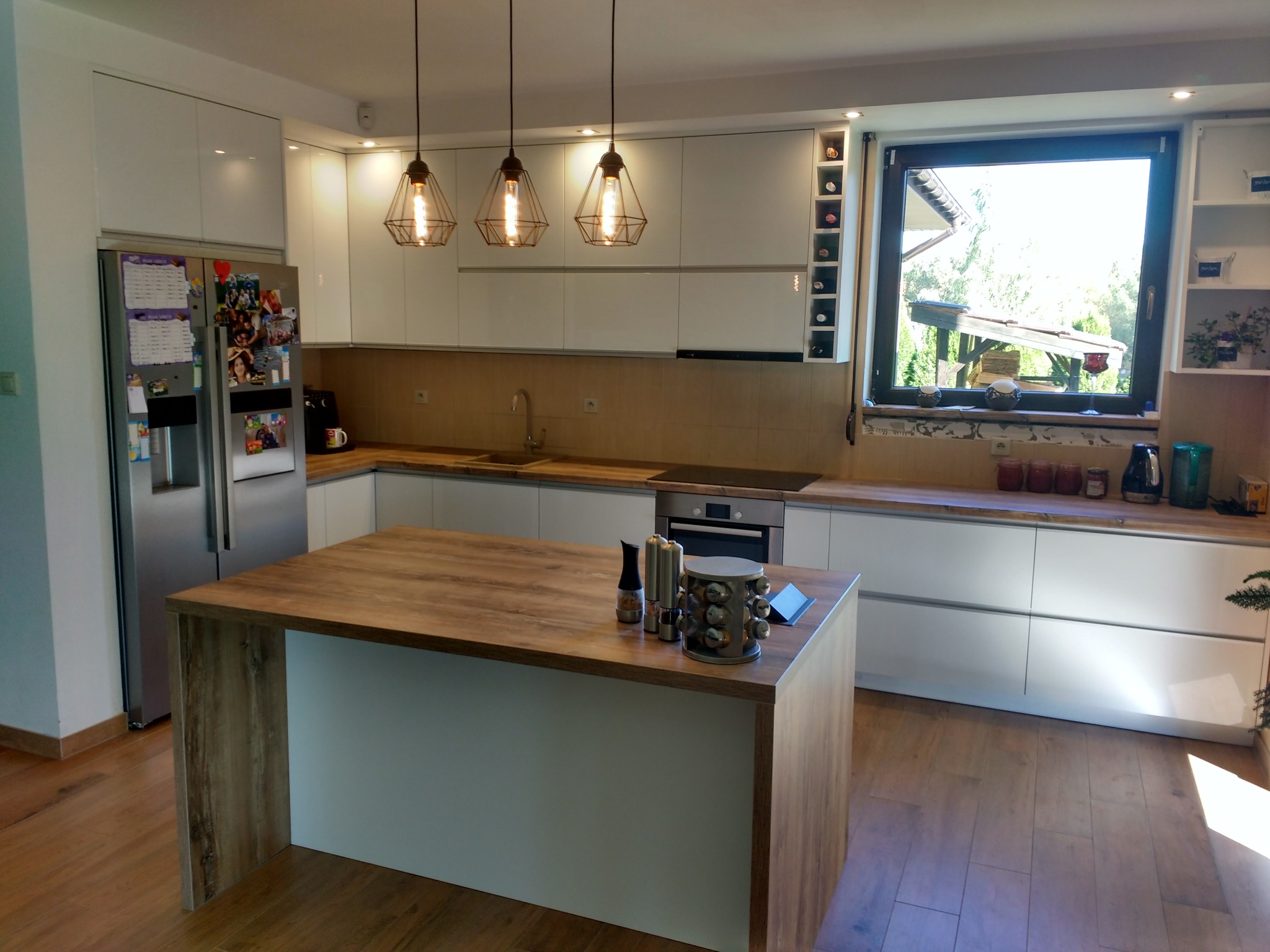 Jasna kuchnia z wyspą, oświetlona trzema lampami z żarówkami Edisona, widok na ogród z okna, minimalistyczne białe szafki i blat z drewna.