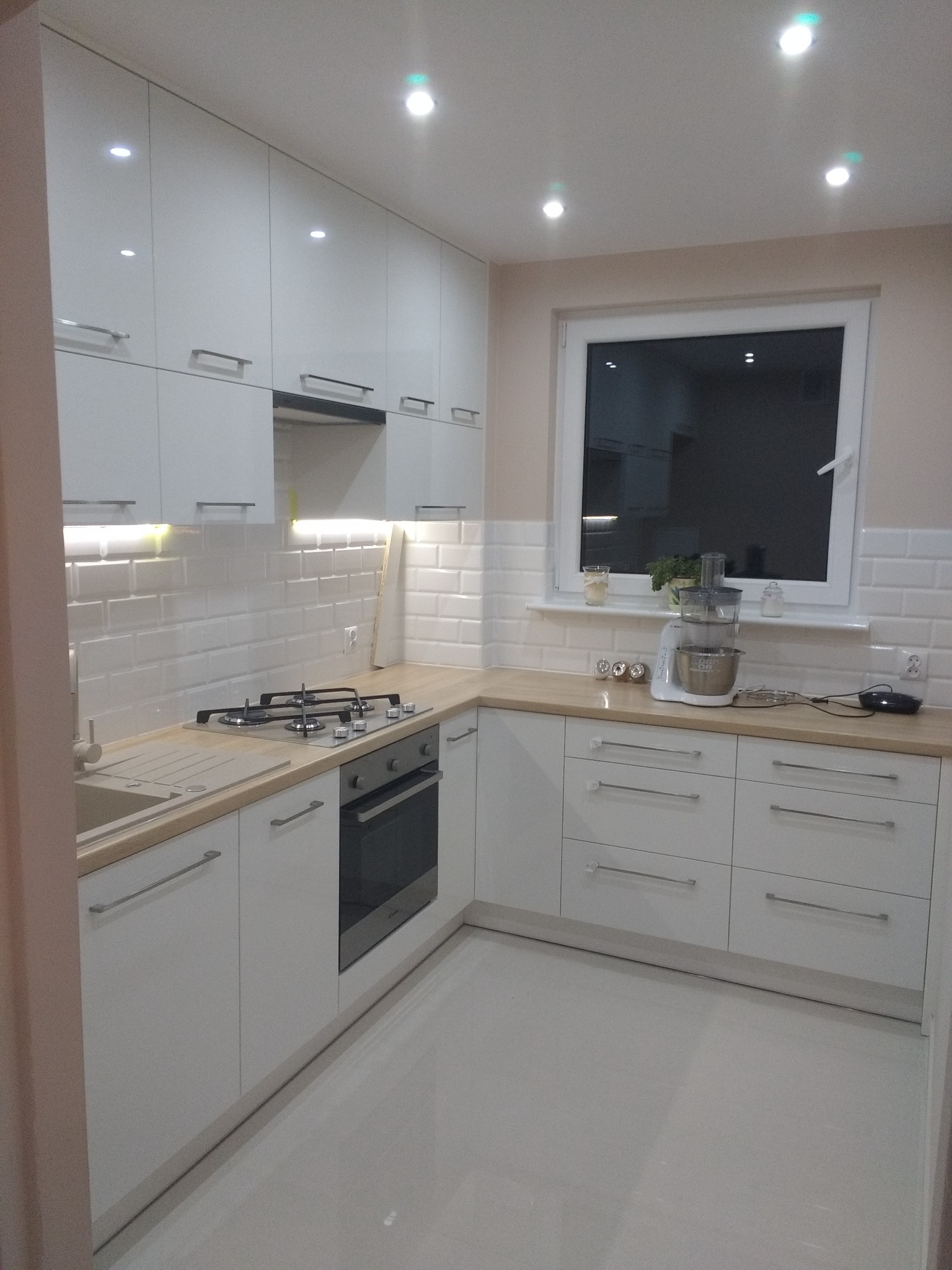 Jasna kuchnia z białymi, błyszczącymi frontami szafek, drewnianym blatem, płytkami typu metro na ścianie oraz sprzętem AGD na blacie pod oknem.