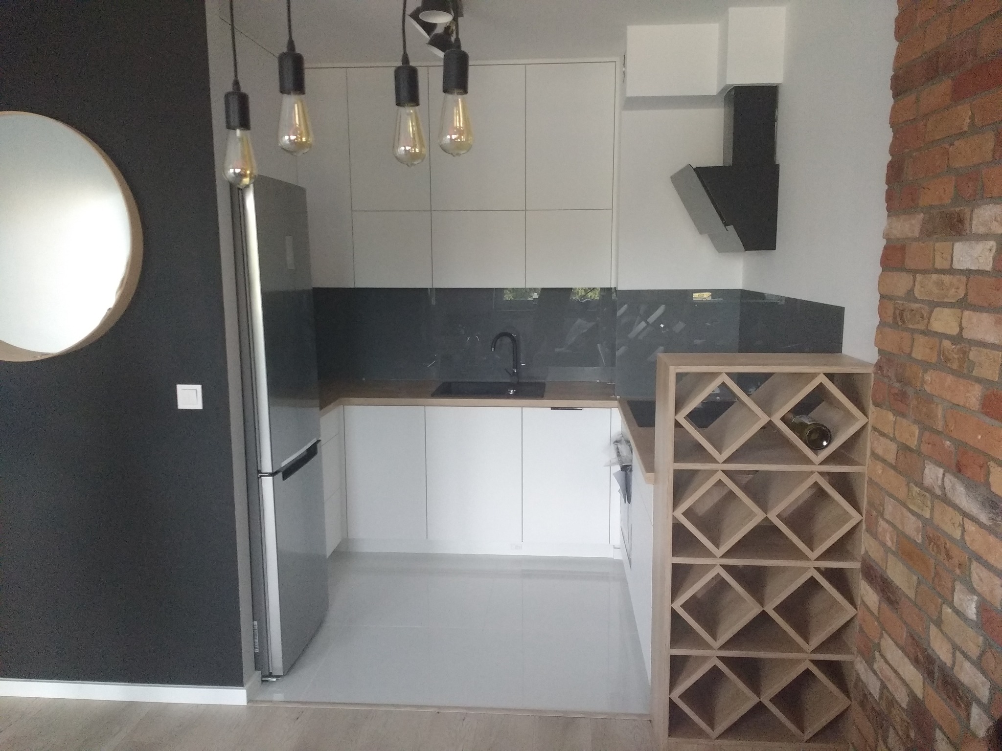 Nowoczesna, minimalistyczna kuchnia w bieli i szarości, z elementami cegły na ścianie, oświetlona wiszącymi żarówkami edisona i wyposażona w regał na wino.