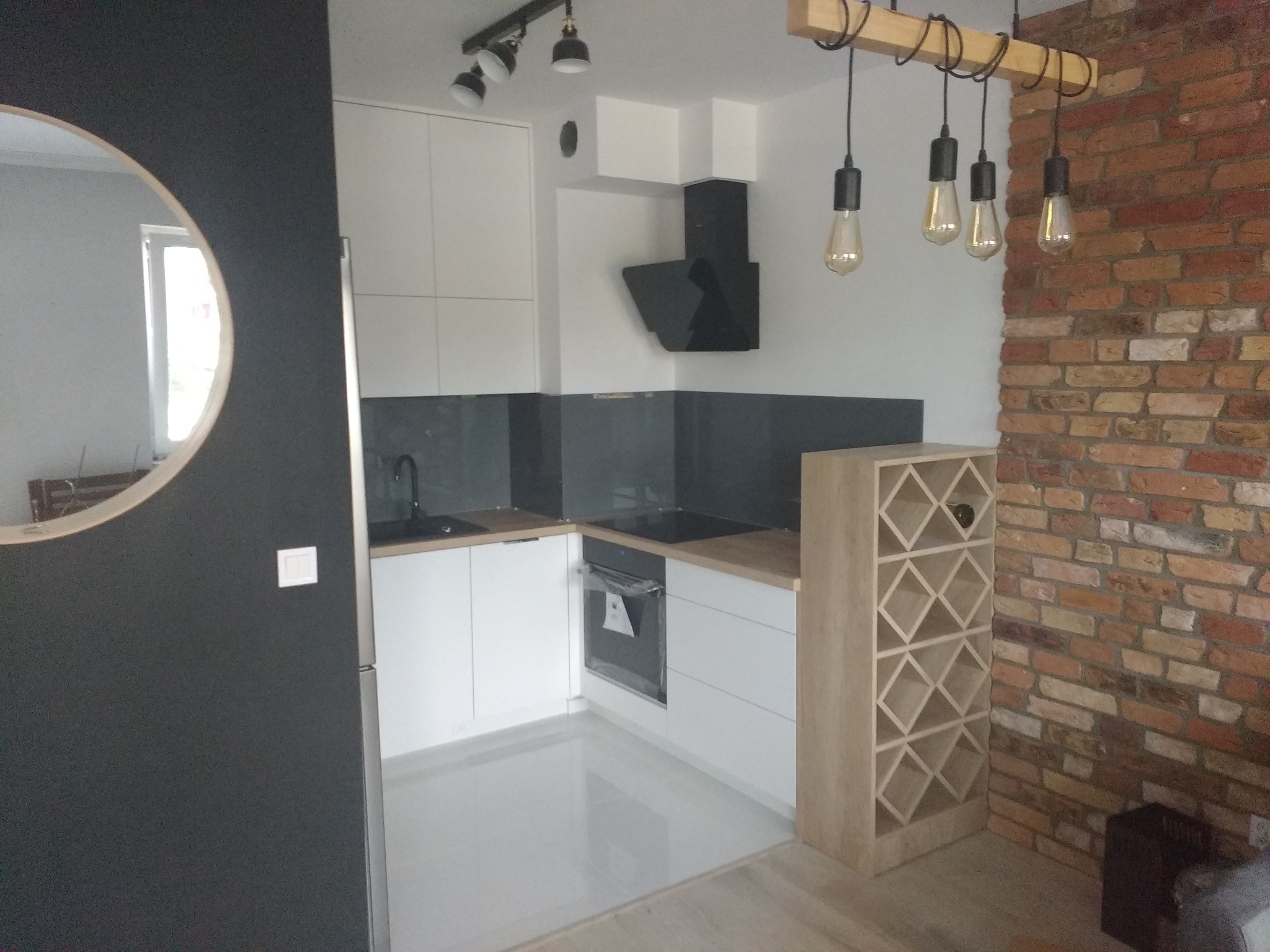 Nowoczesna kuchnia z białymi frontami, szarym backsplashiem, czarnym okapem i ceglaną ścianą z dekoracyjnym oświetleniem żarowym oraz regałem na wino.
