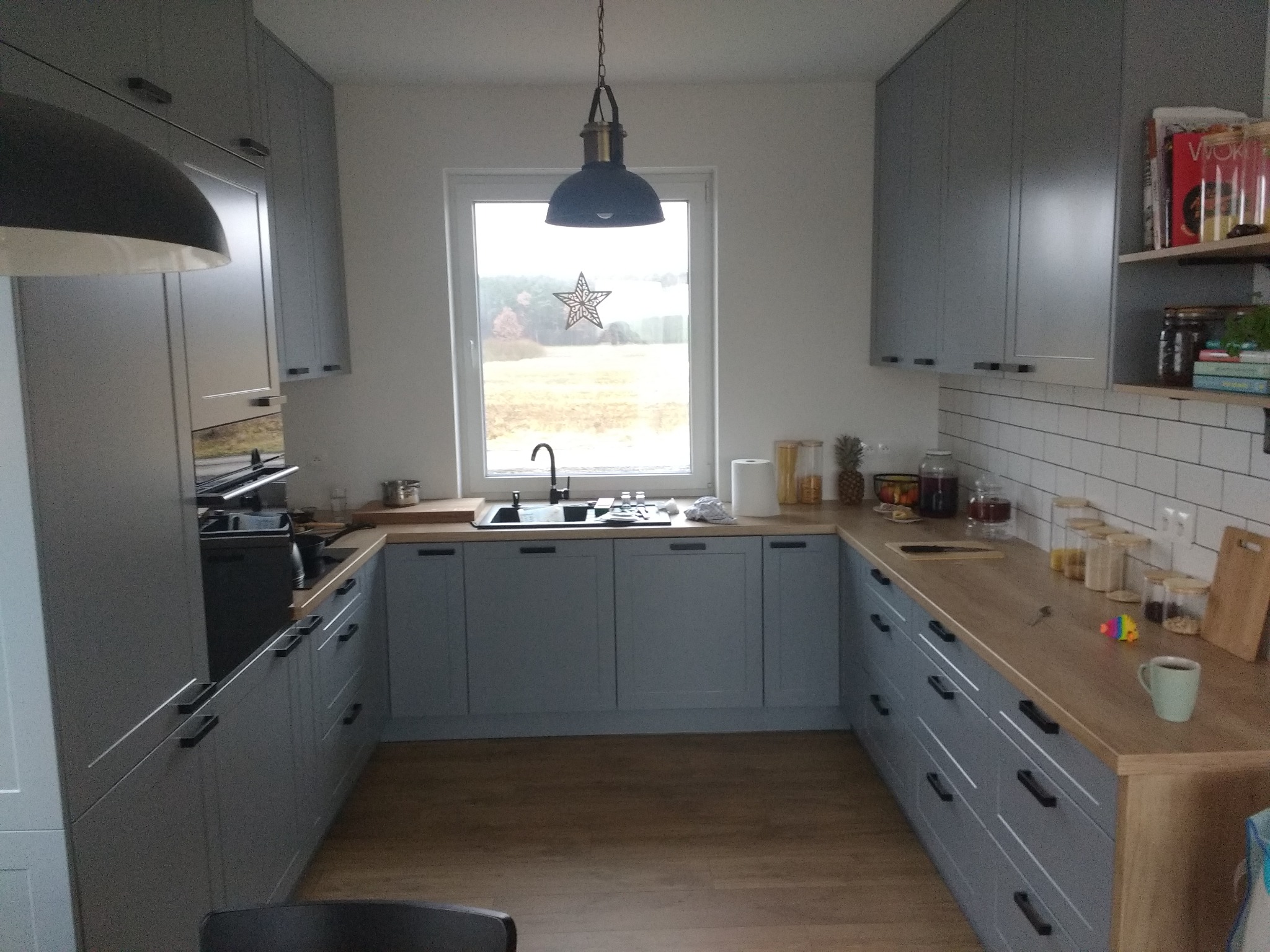 U-kształtna zabudowa kuchenna w odcieniach szarości, z drewnianym blatem i widokiem na krajobraz za oknem, oświetlona wiszącą lampą w stylu industrialnym.