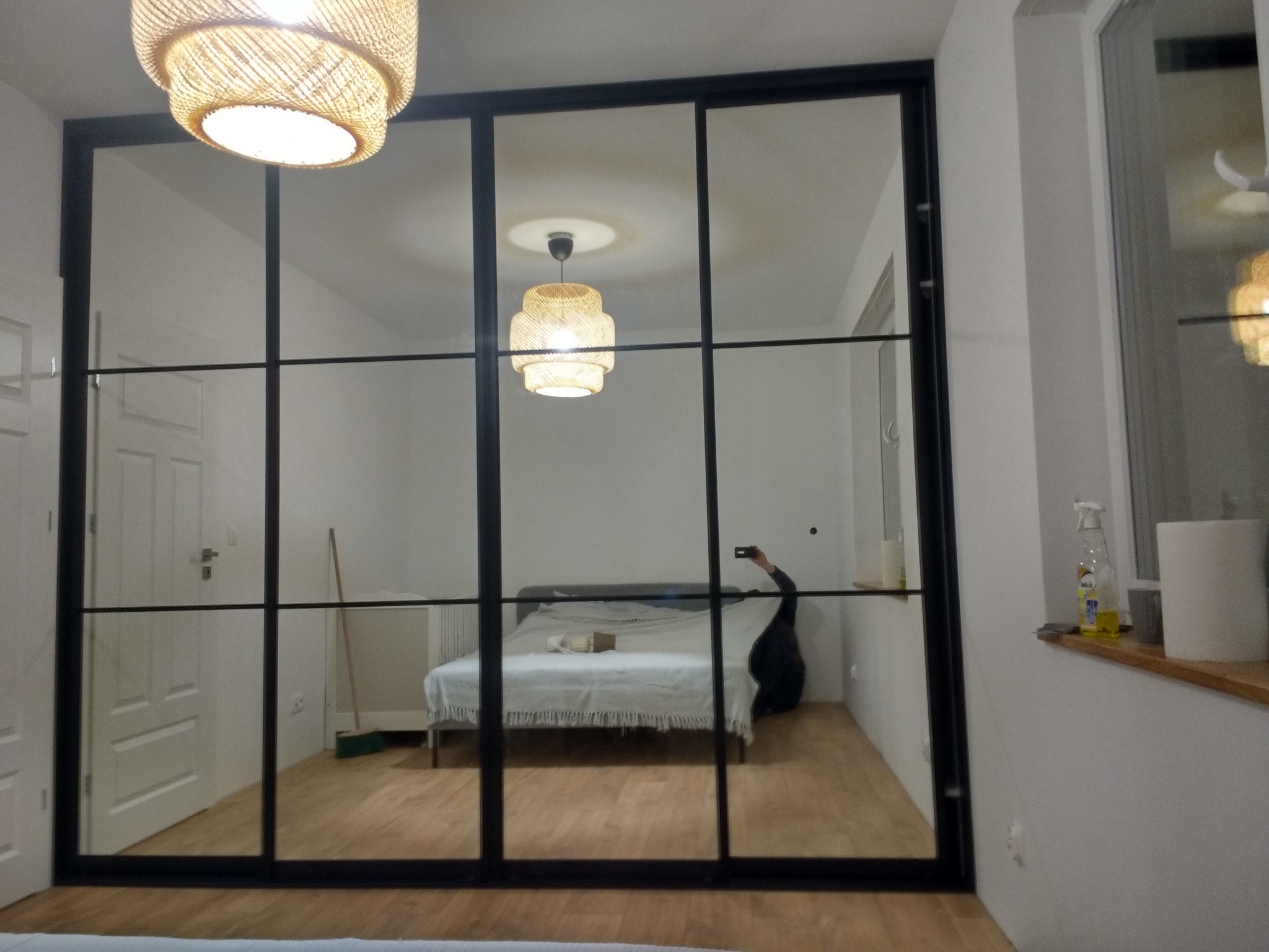 Przesuwne drzwi z czarnymi szprosami, dzielące przestrzeń sypialni, widoczne przez lustrzane tafle odbicie wnętrza z łóżkiem i lampą z abażurem z plecionki.