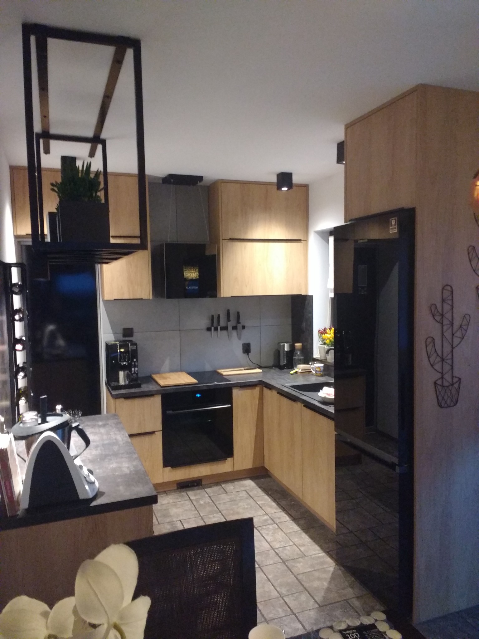 Nowoczesna kuchnia z drewnianymi frontami, grafitowym blatem i czarnymi akcentami, w tym wisząca konstrukcja nad wyspą i dekoracja kaktusa na ścianie.