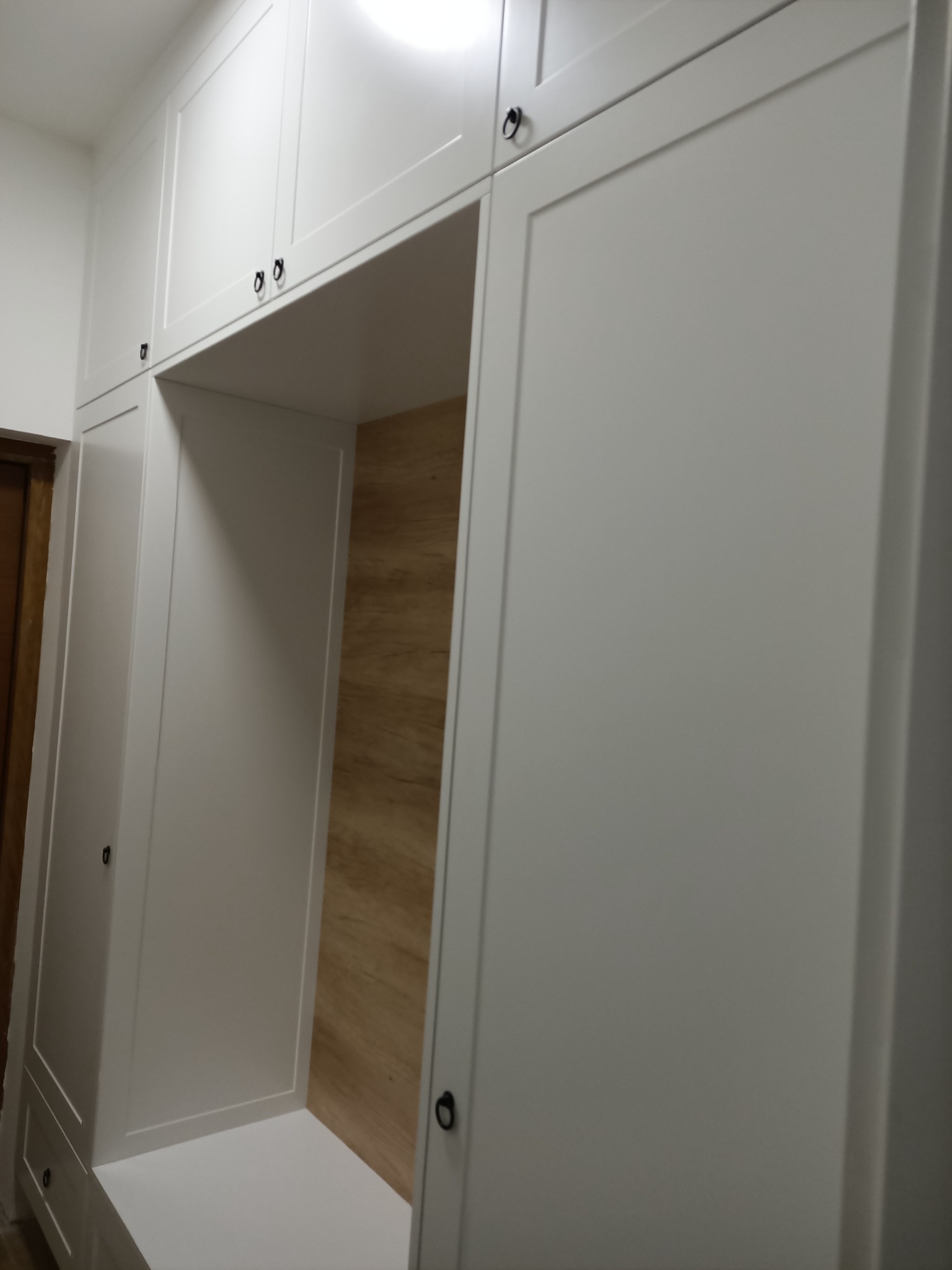 Biała zabudowa meblowa z drewnianym panelem wnęki, czarne okucia, minimalistyczny styl.