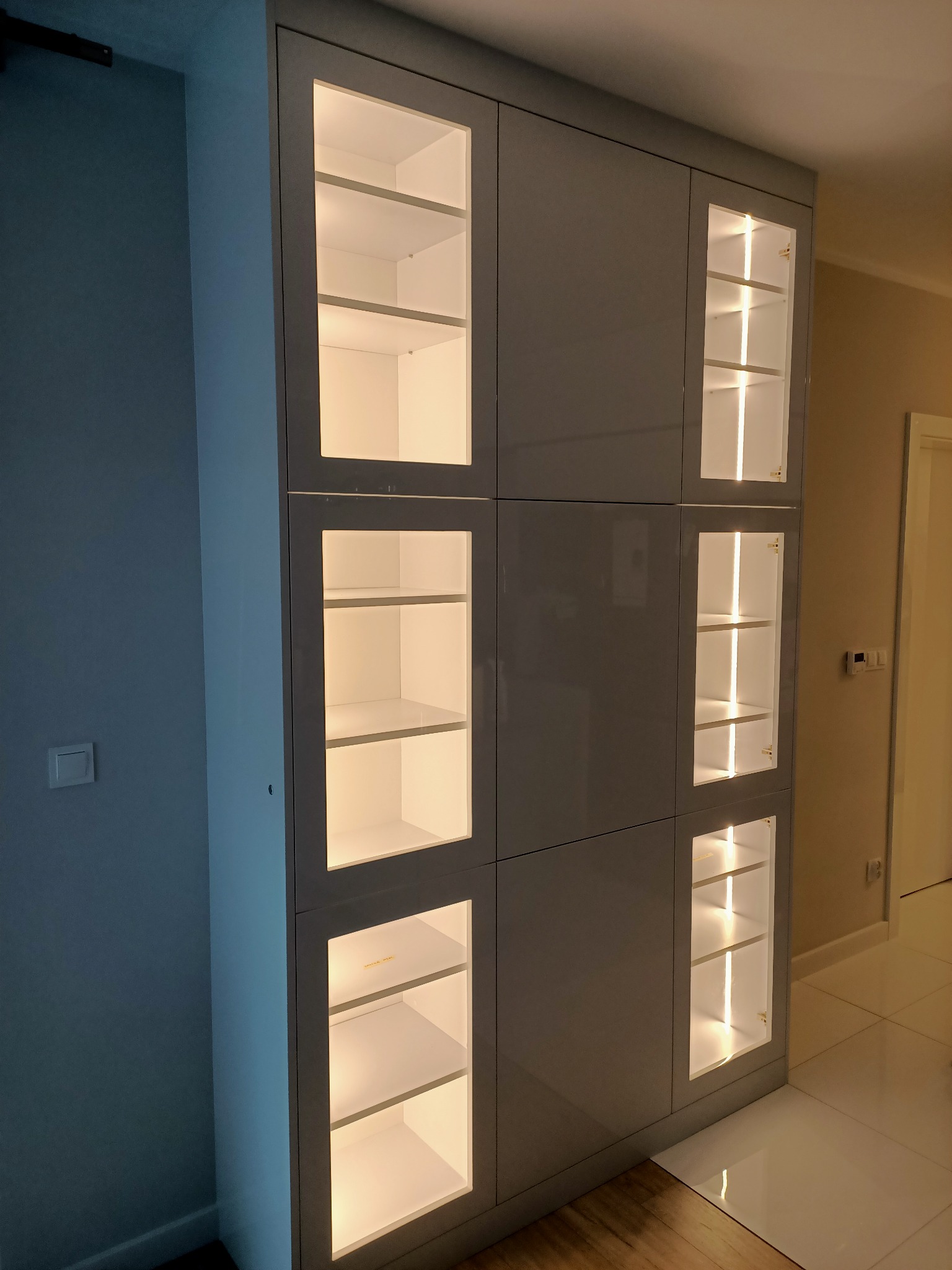 Szara zabudowa meblowa z podświetlanymi półkami za szklanymi frontami, widoczna z perspektywy podłogi, minimalistyczny design.