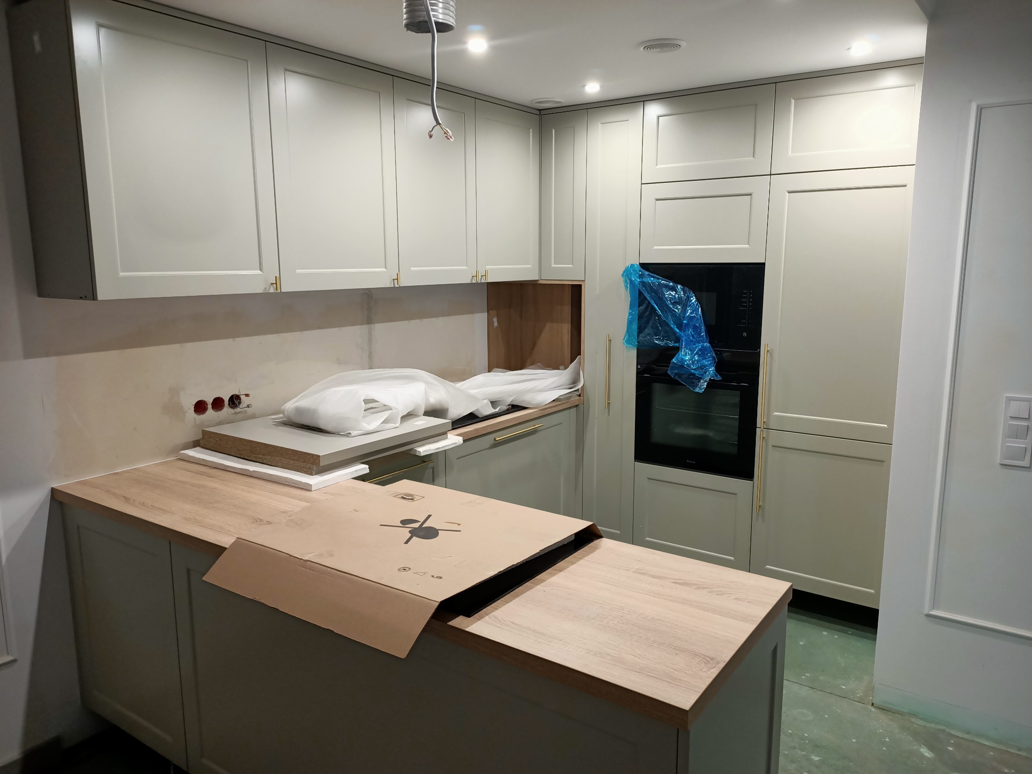 Montaż jasnej kuchni: szafki górne i dolne w odcieniach szarości, drewniany blat roboczy z częściowo zdjętym kartonowym opakowaniem, piekarnik zabezpieczony folią, widoczne gniazdka elektryczne...