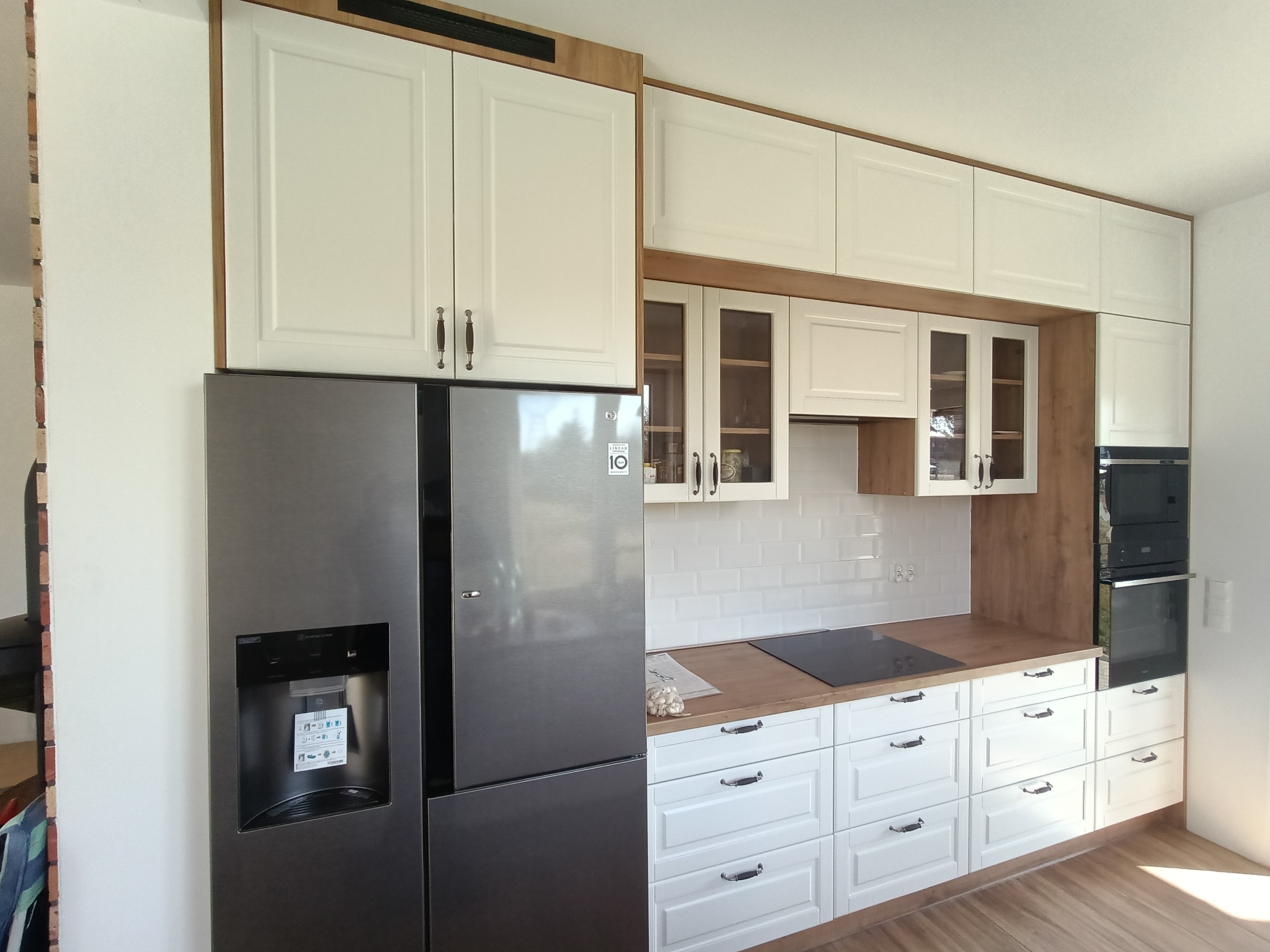 Nowoczesna kuchnia z białymi frontami szafek, drewnianym blatem i obudową, oraz dużą lodówką side-by-side z dystrybutorem wody; widoczne białe płytki na ścianie i płyta indukcyjna.