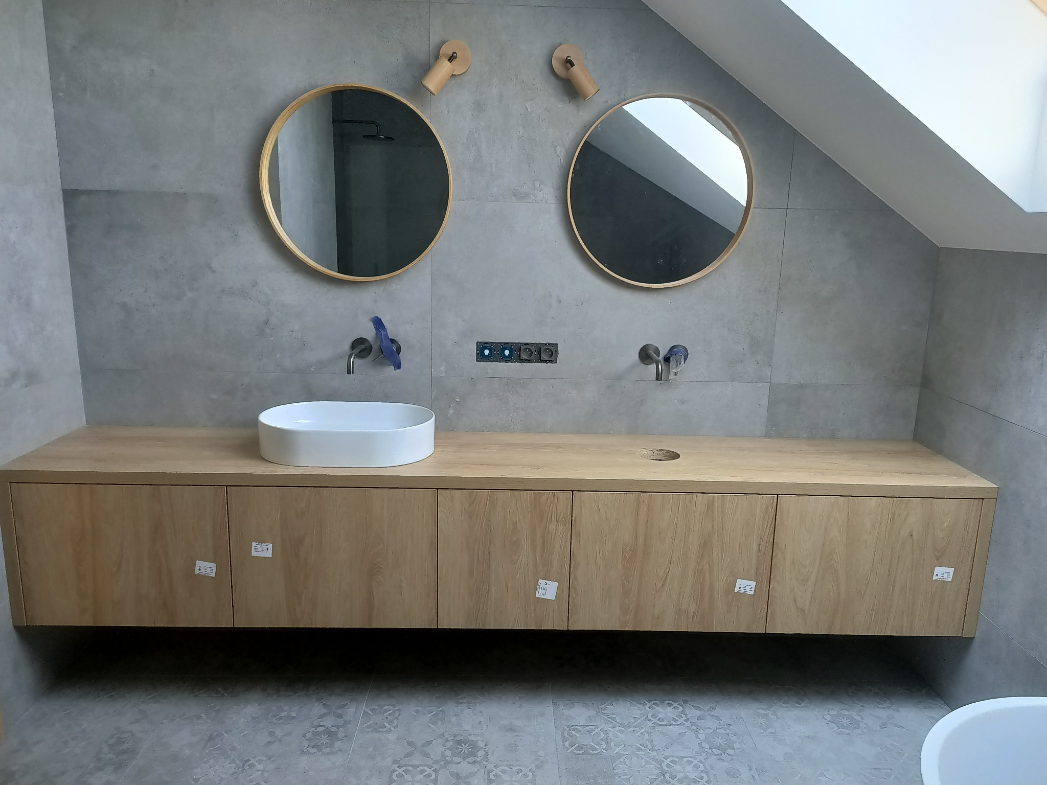 Nowoczesna łazienka na poddaszu z drewnianą szafką pod umywalki, dwoma okrągłymi lustrami z drewnianymi uchwytami i szarą płytką imitującą beton na ścianach i podłodze. Widoczne niebieskie elementy...