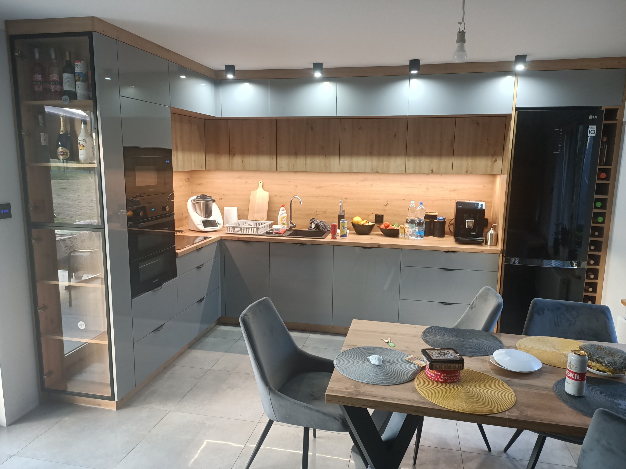 Nowoczesna kuchnia w szarościach i drewnie z wyspą, widok na stół z nakryciami i krzesłami z aksamitną tapicerką, oświetlenie punktowe i wisząca lampa.