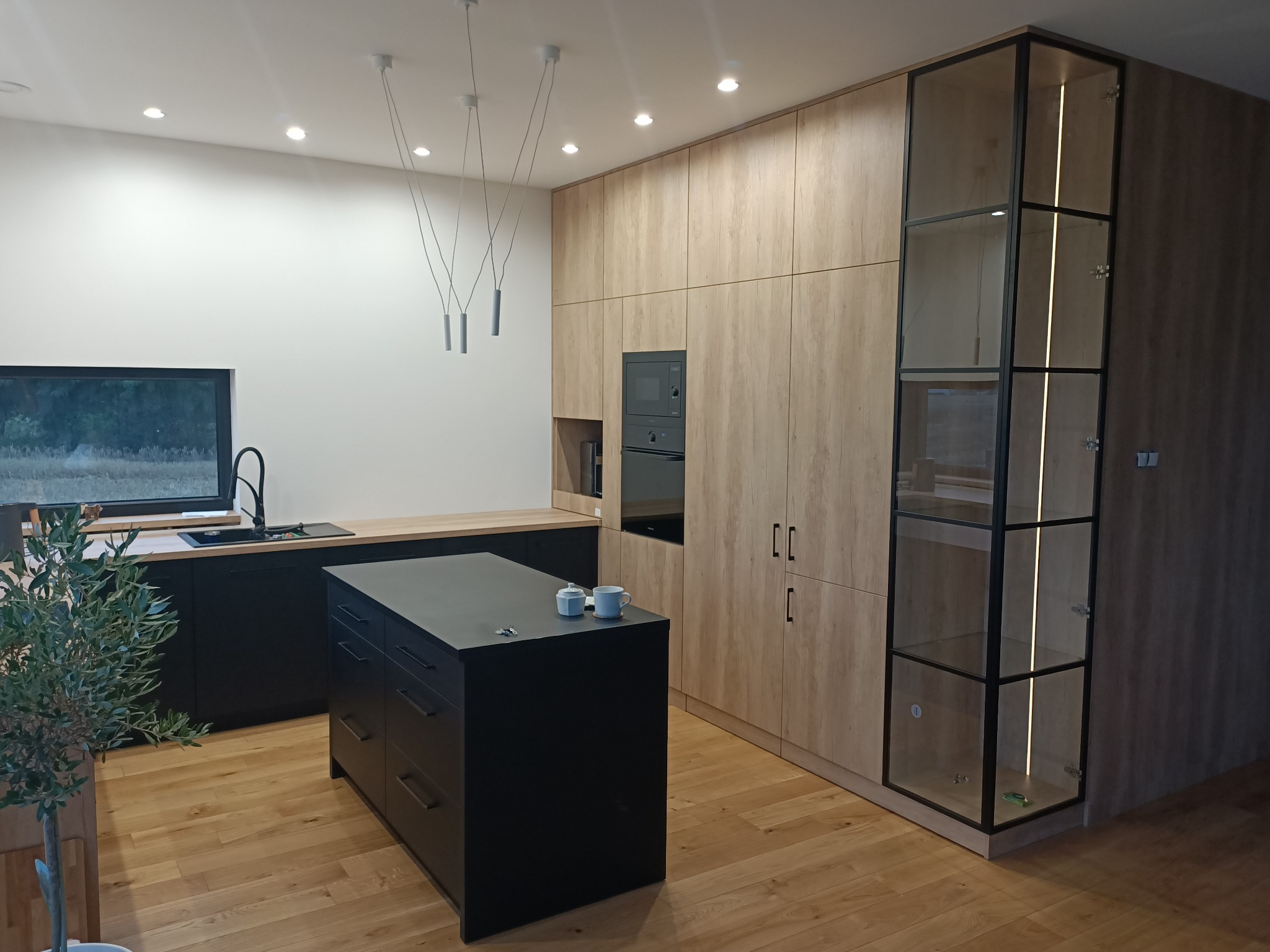 Nowoczesna kuchnia z wyspą i zabudową w odcieniach drewna, czarne fronty, oświetlenie punktowe i wiszące, przeszklona witryna z podświetleniem, drewniana podłoga i roślina doniczkowa w tle okna.