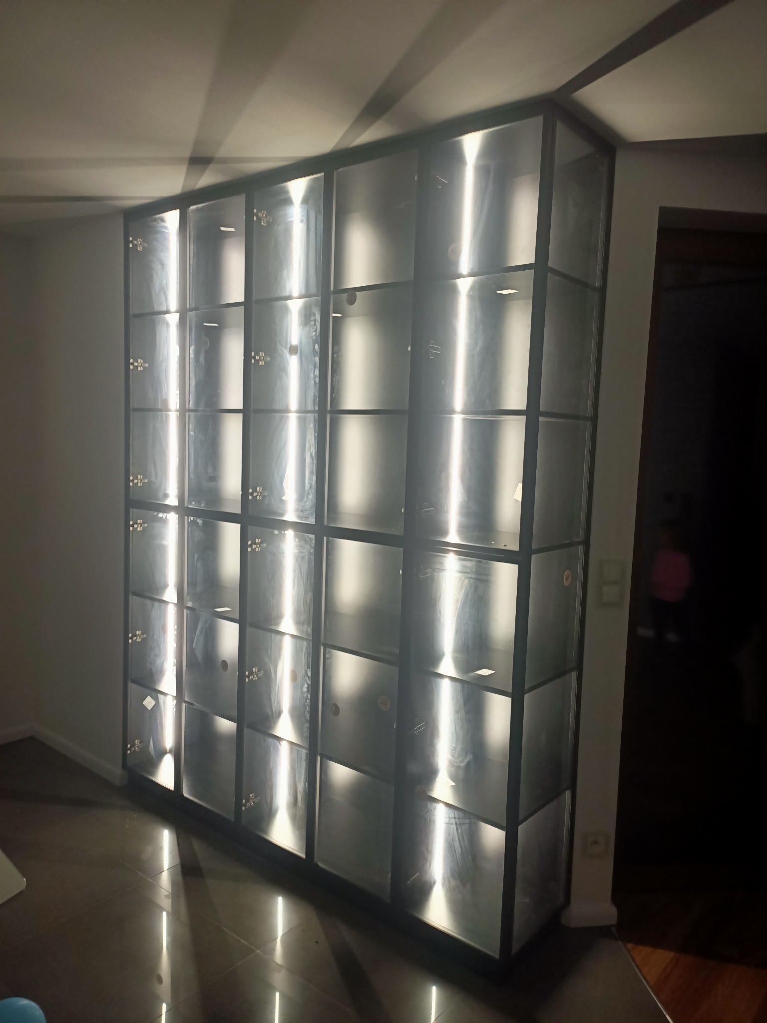 Wysoka, podświetlana od wewnątrz szafa z metalową ramą i szklanymi frontami, podzielona na liczne kwadratowe przegródki, odbijająca światło na podłodze.
