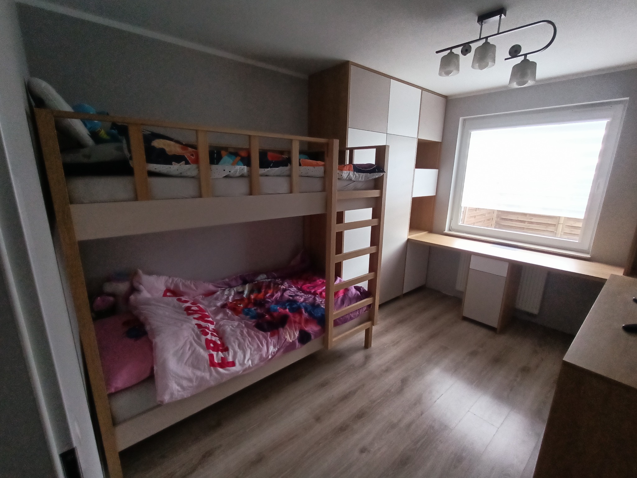 Pokój dziecięcy z drewnianym łóżkiem piętrowym, szafą w zabudowie z białymi frontami i biurkiem pod oknem, oświetlonym lampą sufitową z trzema kloszami.