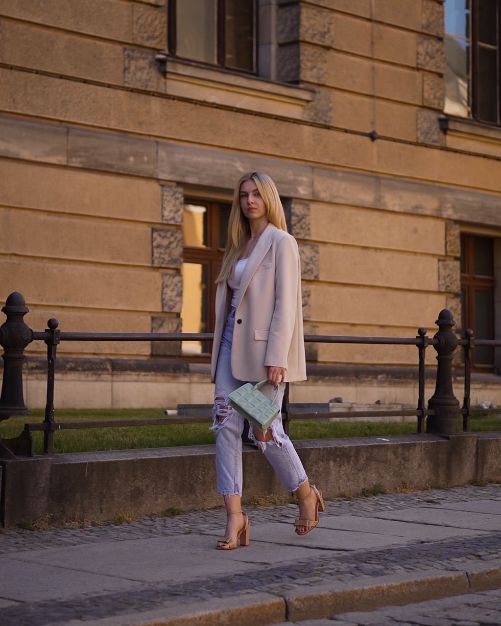 Blondwłosa kobieta w beżowej marynarce, podartych jeansach i sandałach na obcasie, trzymająca małą, kwadratową torebkę, spaceruje po chodniku na tle budynku z kamienia.