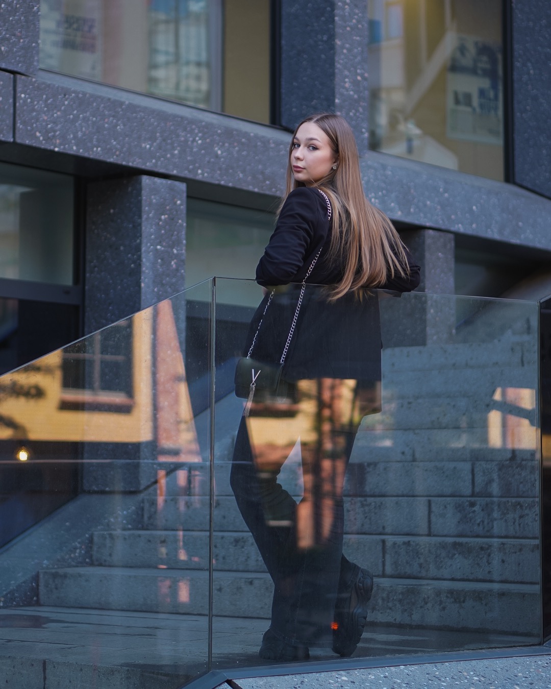 Młoda kobieta w czarnym garniturze, spoglądająca przez ramię na tle nowoczesnej architektury miejskiej i szklanej balustrady, z widocznym odbiciem schodów i budynku.