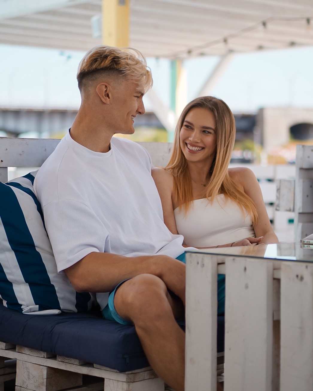 Uśmiechnięta para, blondyn i blondynka, siedząca obok siebie na paletach pomalowanych na biało, z niebieskimi poduszkami w paski, przy szklanym stoliku, patrząc na siebie z czułością.