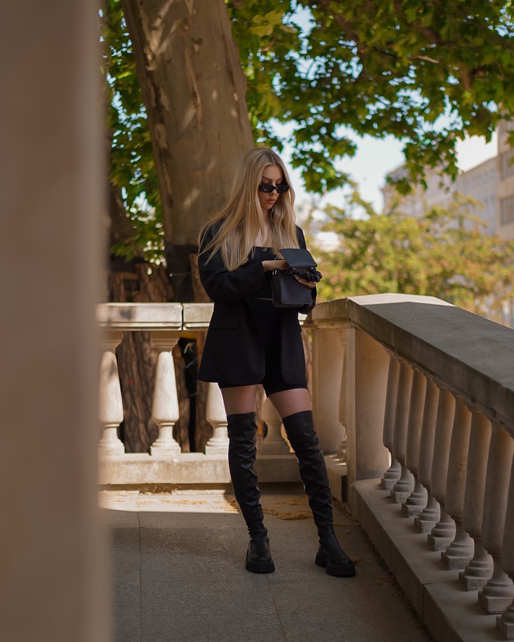 Stylowa blondynka w czarnym komplecie (żakiet, szorty, kozaki za kolano) i okularach przeciwsłonecznych, trzymająca czarną torebkę na tle balustrady i drzewa.
