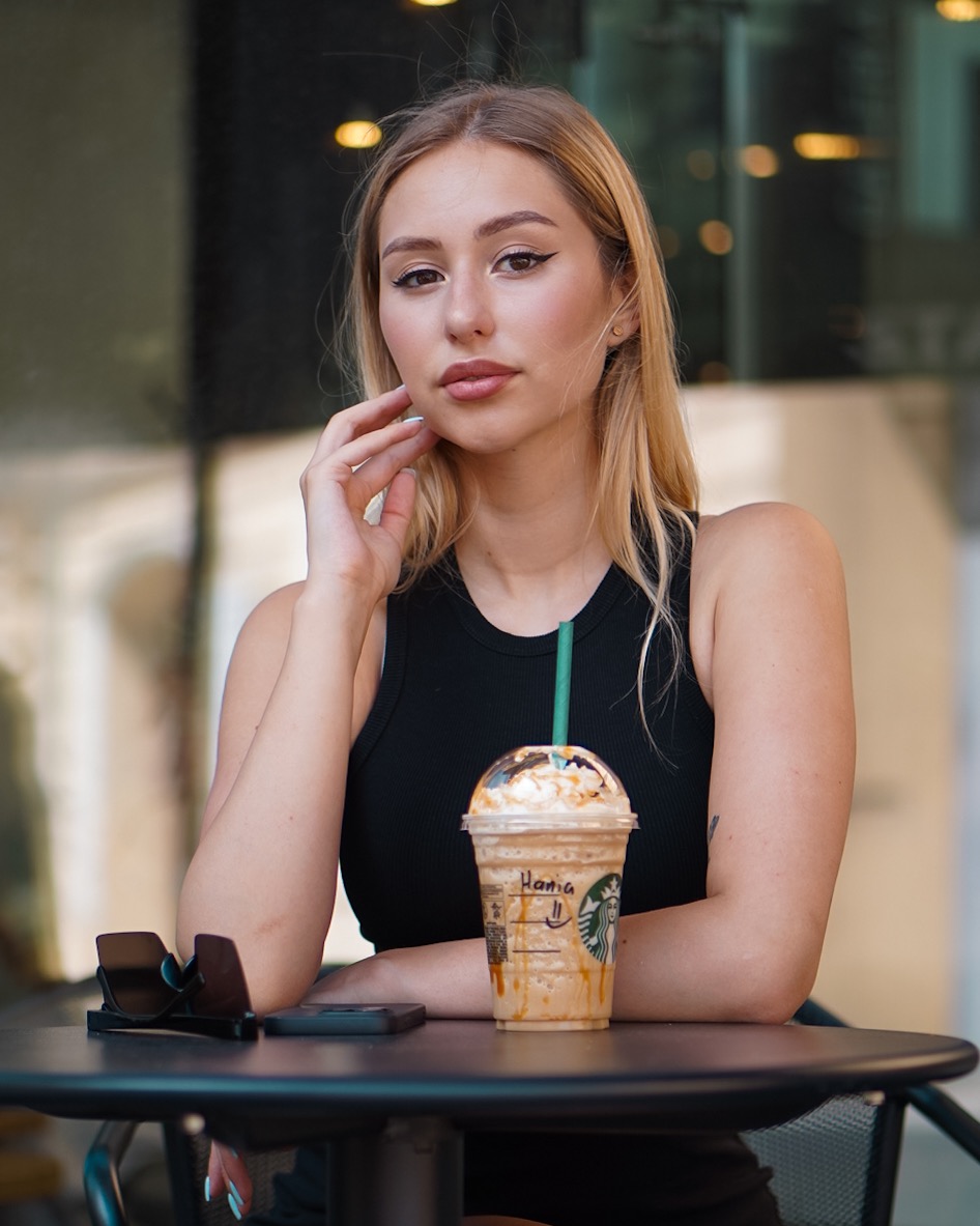 Portret młodej kobiety z blond włosami, siedzącej przy stoliku z napojem Starbucks, z okularami i telefonem obok.