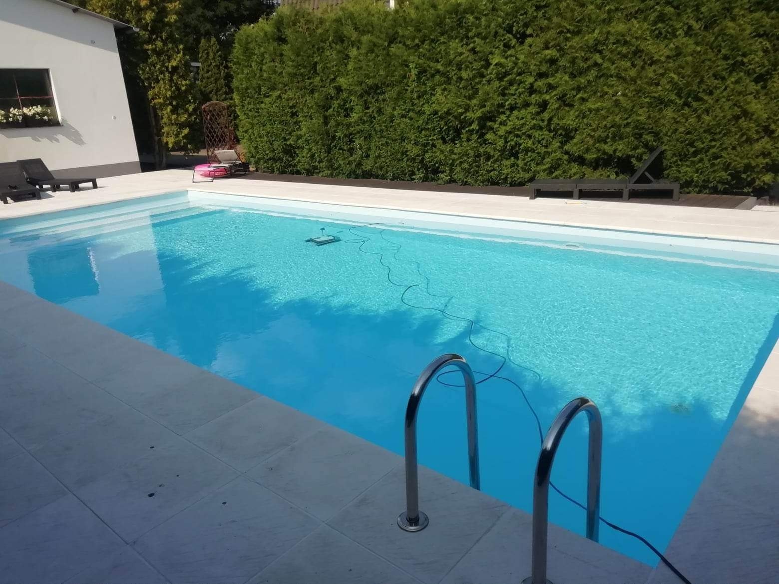 Błękitny basen ogrodowy z chromowanymi schodkami, otoczony jasnymi płytkami i zielonym żywopłotem, z widocznym robotem czyszczącym na dnie.