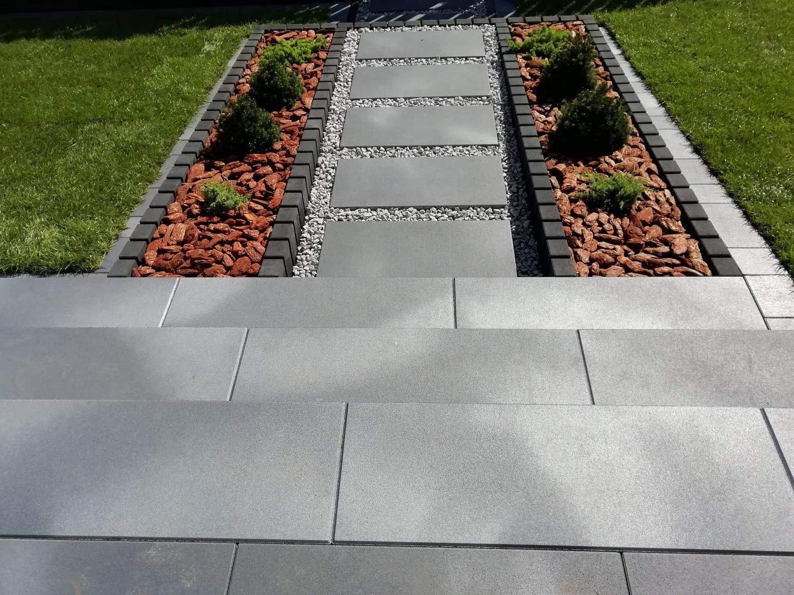 Schody z szarych płyt prowadzące do ścieżki z prostokątnych płyt, obramowanej szarym i czarnym obrzeżem, z rabatami wypełnionymi korą i niskimi, zielonymi krzewami.