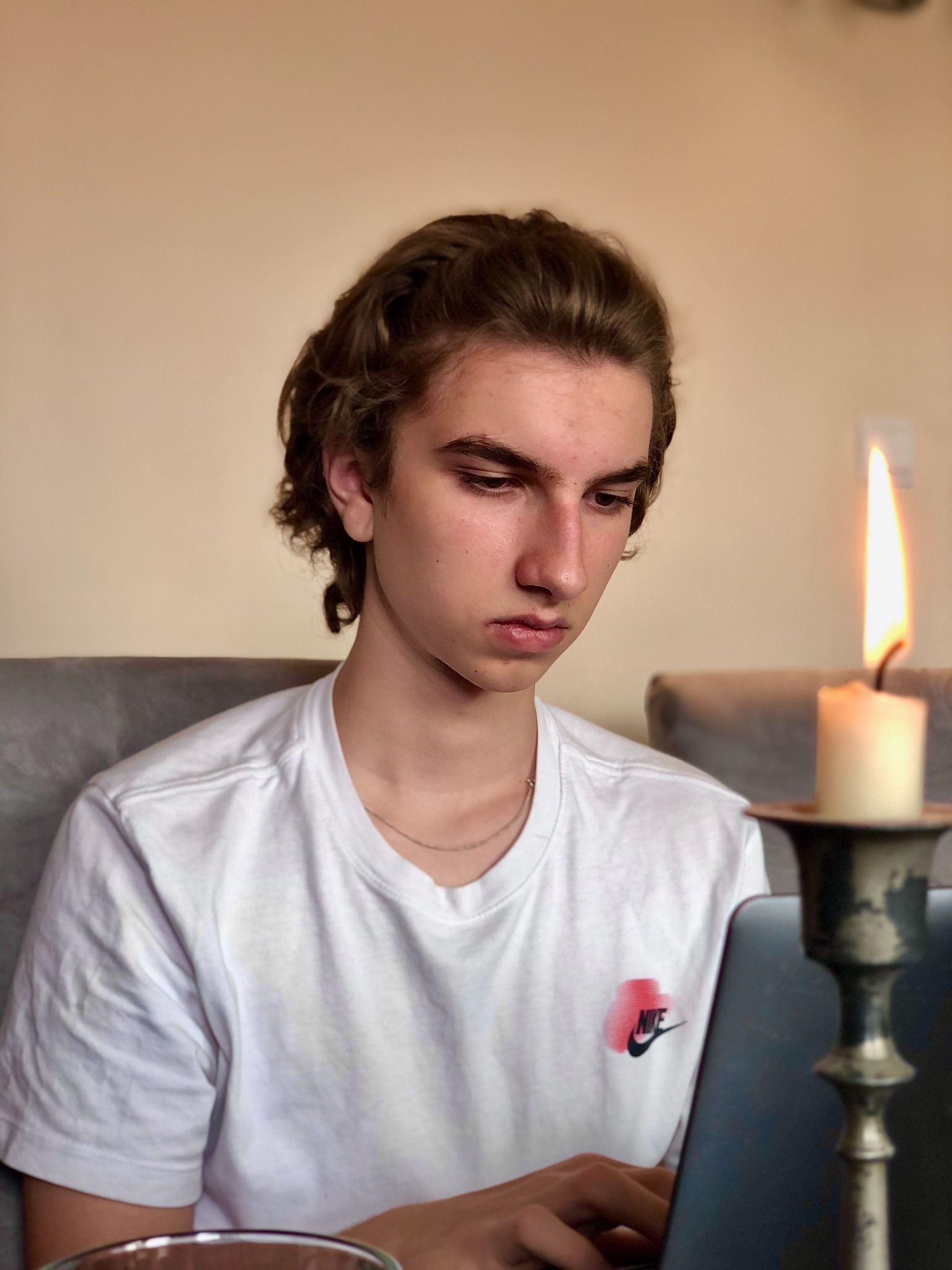 Młody mężczyzna skupiony na pracy przy laptopie, oświetlonym płomieniem świecy w metalowym świeczniku, z profilu widoczny napis Nike na białej koszulce.