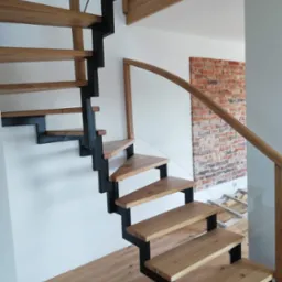 Drewniane schody z czarną, metalową konstrukcją wspornikową, zamontowane w nowoczesnym wnętrzu z fragmentem ściany z cegły.