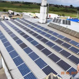 Rząd paneli słonecznych na dachu budynku przemysłowego z widocznym fragmentem drabiny i ręką, tło stanowią pola i zabudowania.