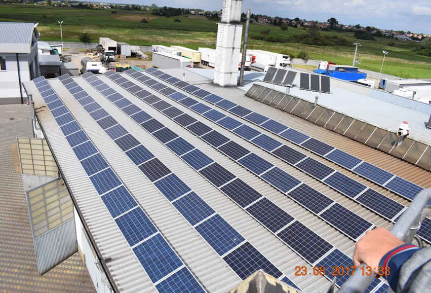 Rząd paneli słonecznych na dachu budynku przemysłowego z widocznym fragmentem drabiny i ręką, tło stanowią pola i zabudowania.