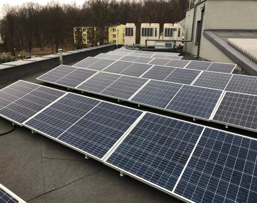Rząd paneli słonecznych na płaskim dachu budynku, z widocznymi klimatyzatorami na dachu w tle i drzewami w oddali.