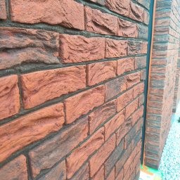 SamRemo - Fragment elewacji budynku z czerwonej cegły klinkierowej o wyraźnej fakturze, widoczny narożnik budynku i fragment obsypanego kamieniami podłoża z zieloną rośliną.