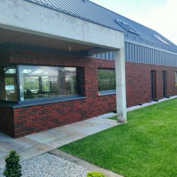 SamRemo - Nowoczesny dom jednorodzinny z elewacją z czerwonej cegły klinkierowej, metalowym dachem i dużymi oknami; na zewnątrz trawnik i chodnik z płyt.