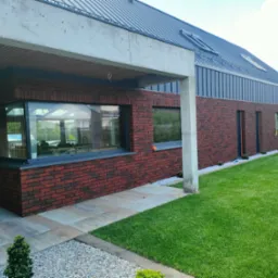 Nowoczesny dom jednorodzinny z elewacją z czerwonej cegły klinkierowej, metalowym dachem i dużymi oknami; na zewnątrz trawnik i chodnik z płyt.