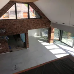 Przestronne wnętrze w trakcie prac wykończeniowych, widok z góry na nową, jasnoszarą podłogę z płytek, ściany częściowo wykończone czerwoną cegłą, widoczne okna z widokiem na rusztowania...