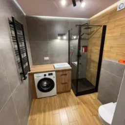 Nowoczesna łazienka z kabiną prysznicową typu walk-in, umywalką nablatową na drewnianej szafce, pralką i grzejnikiem drabinkowym na szarej, płytkowej ścianie.