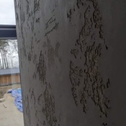 SamRemo - Szara ściana z dekoracyjną, strukturalną fakturą, widoczne okno z widokiem na zewnątrz i niebieska plandeka na podwórku.