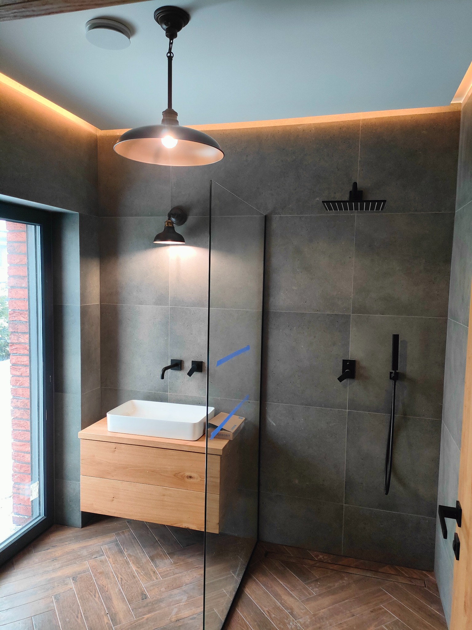 Nowoczesna łazienka z szarymi płytkami, drewnianą podłogą ułożoną w jodełkę, wiszącą szafką z umywalką, czarną armaturą i oświetleniem LED.