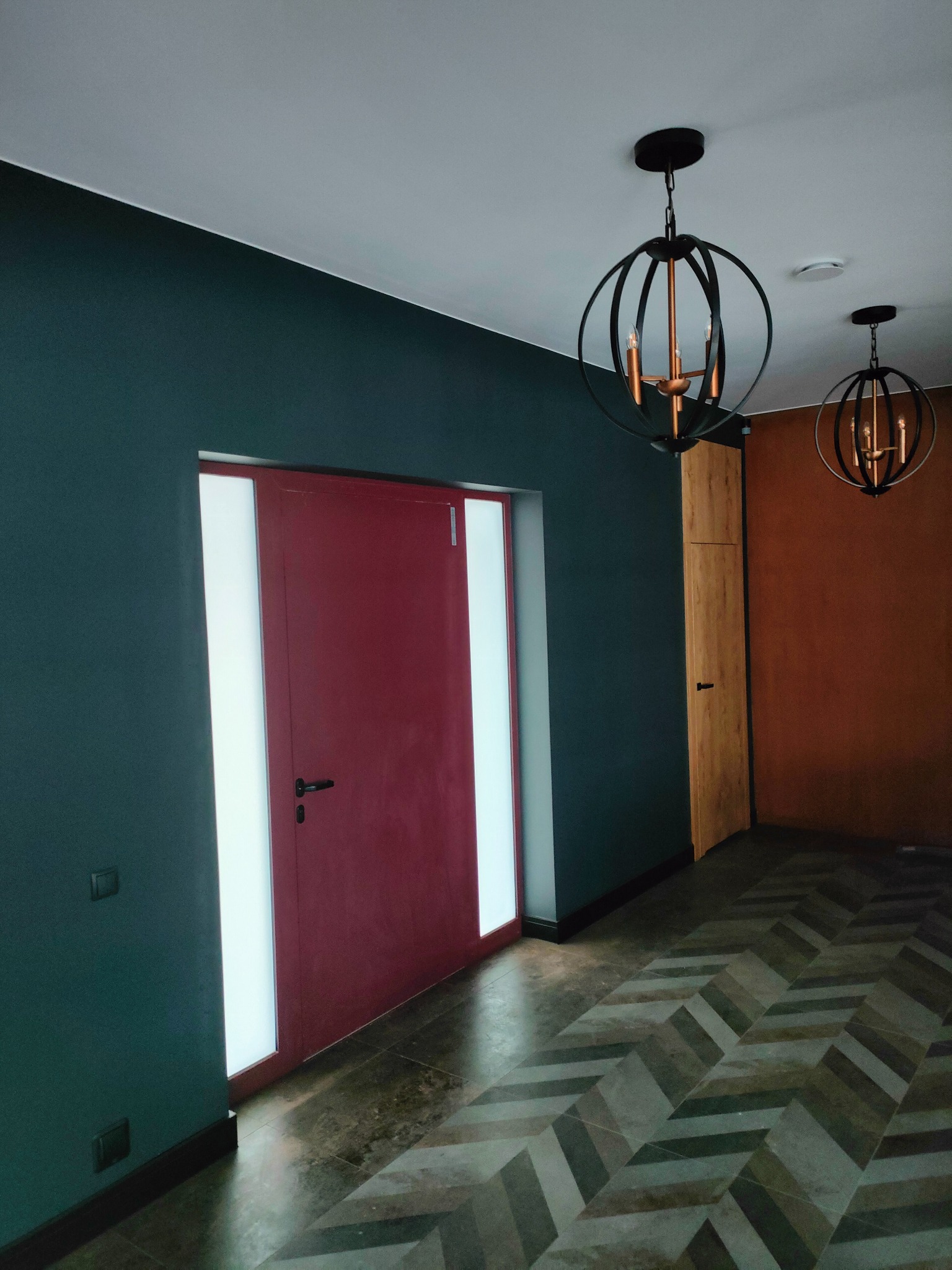 Nowoczesne wnętrze: ciemnozielona ściana, bordowe drzwi z przeszkleniami, parkiet w jodełkę i okrągłe lampy wiszące z metalowymi obręczami.