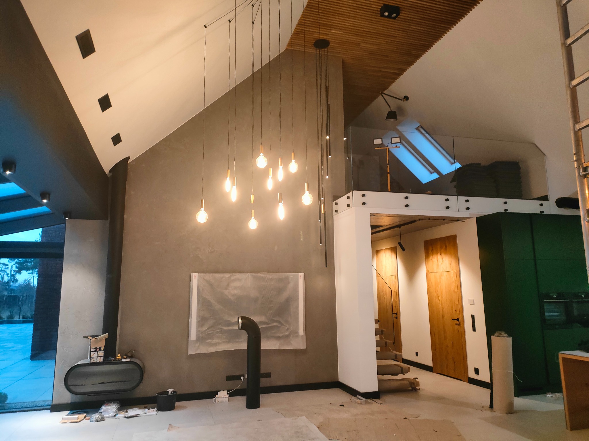 Wnętrze w trakcie prac wykończeniowych: szpachlowana ściana z osłoniętym miejscem na obraz, nowoczesny kominek wolnostojący, wisząca lampa z wieloma żarówkami, drewniany sufit, schody i drzwi w tle.