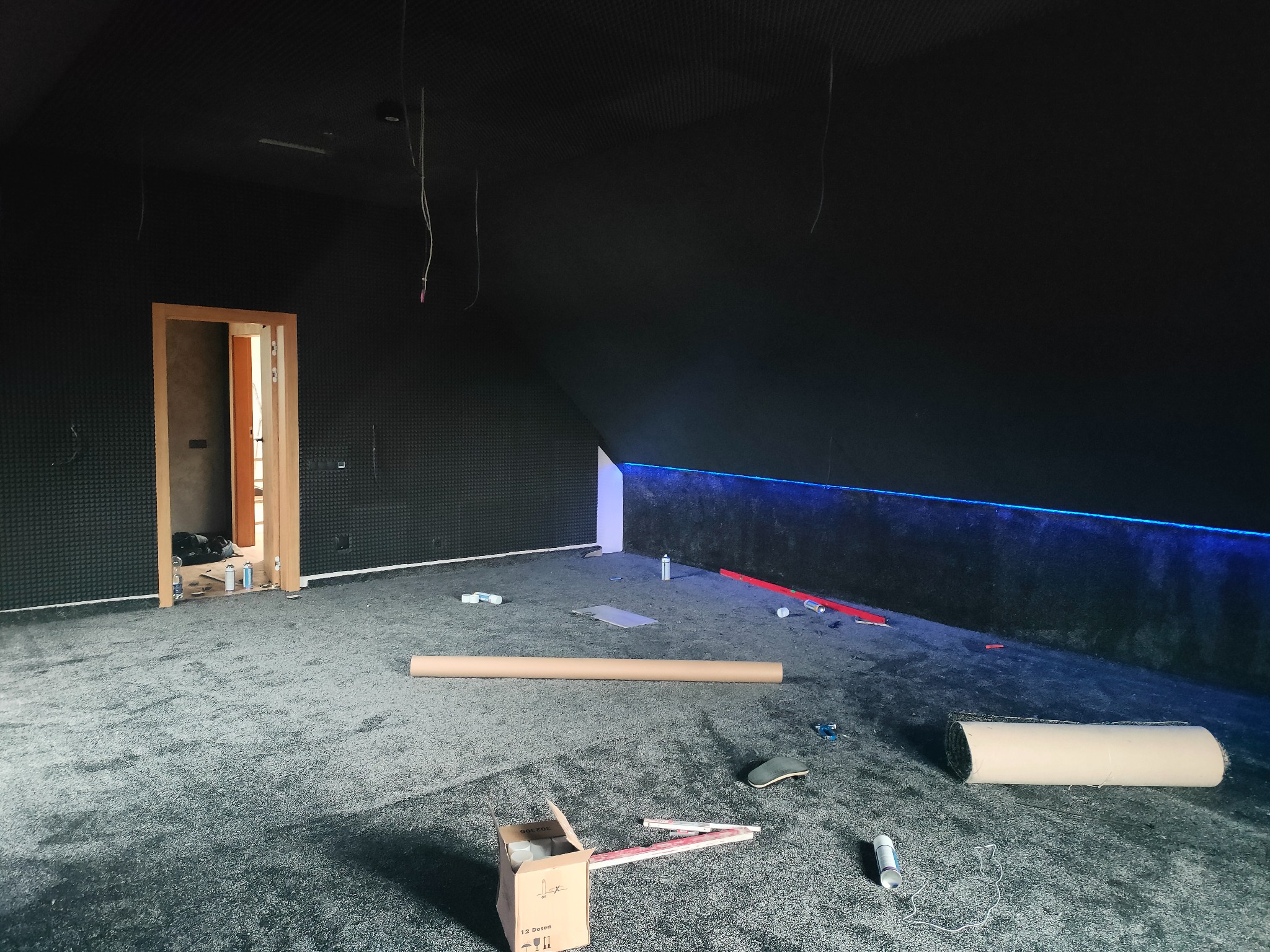 Pokój w trakcie prac wykończeniowych: czarne ściany z panelami akustycznymi, szara wykładzina, otwarte drzwi, widoczne narzędzia i materiały budowlane, niebieskie oświetlenie LED na ścianie.