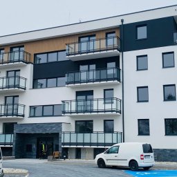 Usługi Inżynierskie RTK-BUD Robert Kmiecik - Nowoczesny budynek wielorodzinny z balkonami, wykończony w bieli i czerni, z elementami drewna. Na pierwszym planie zaparkowane busy.