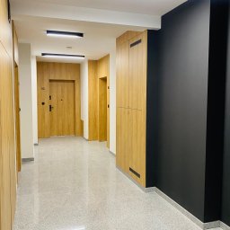Usługi Inżynierskie RTK-BUD Robert Kmiecik - Korytarz budynku wielorodzinnego z drewnianymi drzwiami i szafami w zabudowie. Podłoga z lastryko, czarna ściana, oświetlenie liniowe LED. Nowoczesne wykończenie wnętrz.