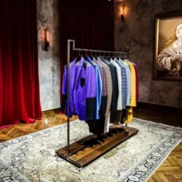 Manekin w marynarce w pepitkę obok wieszaka z kolorowymi marynarkami, na tle czerwonych zasłon i obrazu w złotej ramie.