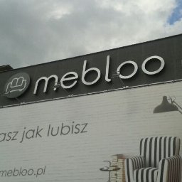 Pracownia Reklamy Plastica Tabernacka Sp. j - Elewacja budynku z szyldem 'mebloo' i billboardem reklamowym przedstawiającym fotel w paski, lampę podłogową i hasło 'Urządzasz jak lubisz'.