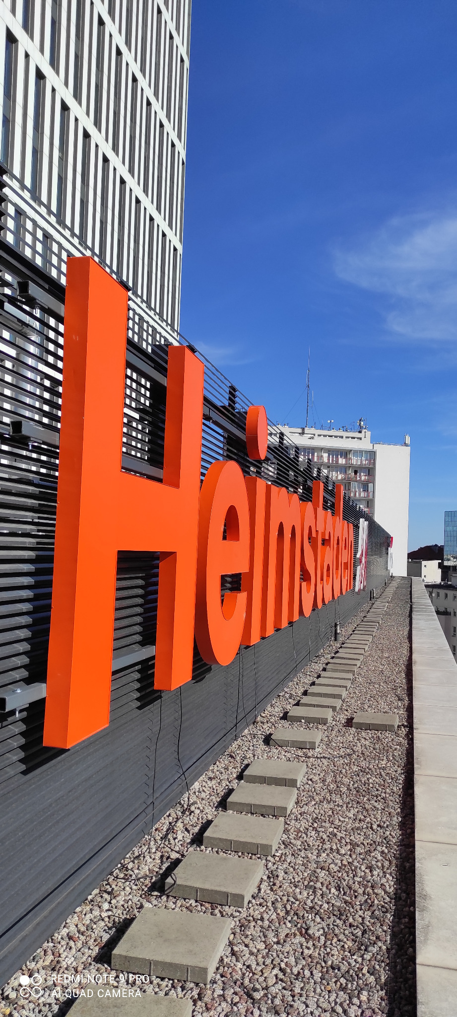 Pomarańczowe, przestrzenne litery tworzące napis 'Heimstaden' na dachu budynku z szarymi panelami, widok z dołu w słoneczny dzień.