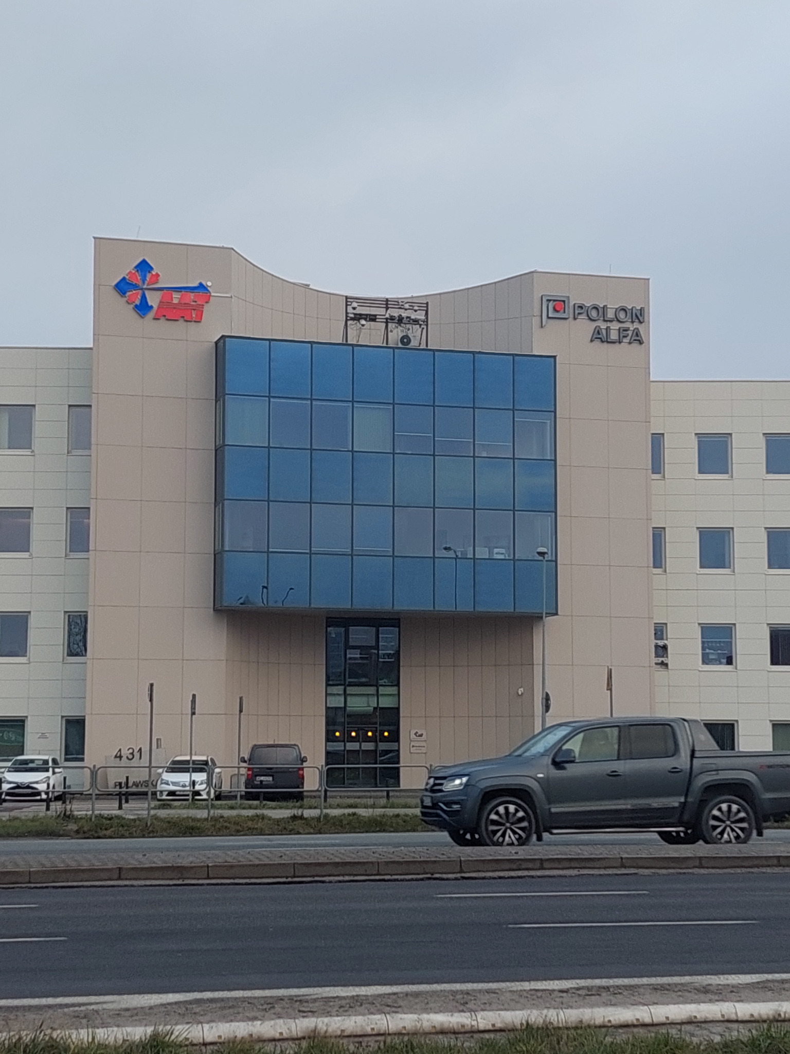 Budynek biurowy z logo firm na fasadzie w Warszawie, widoczny z ulicy z przejeżdżającym samochodem.