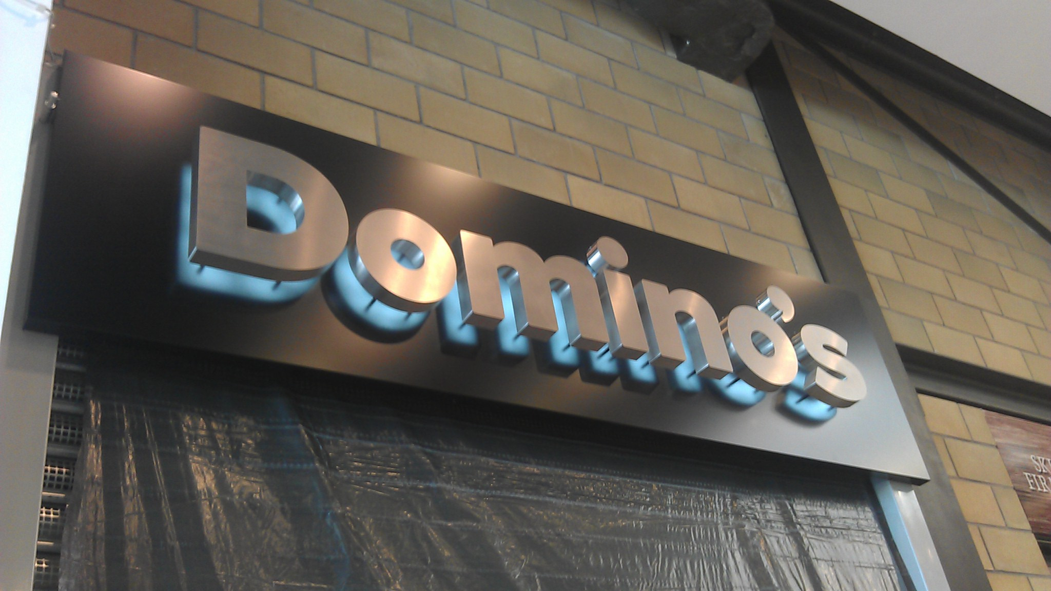 Metalowe litery przestrzenne z podświetleniem tworzące szyld Domino's zamontowany na ścianie z cegieł, z widoczną folią ochronną na dole.