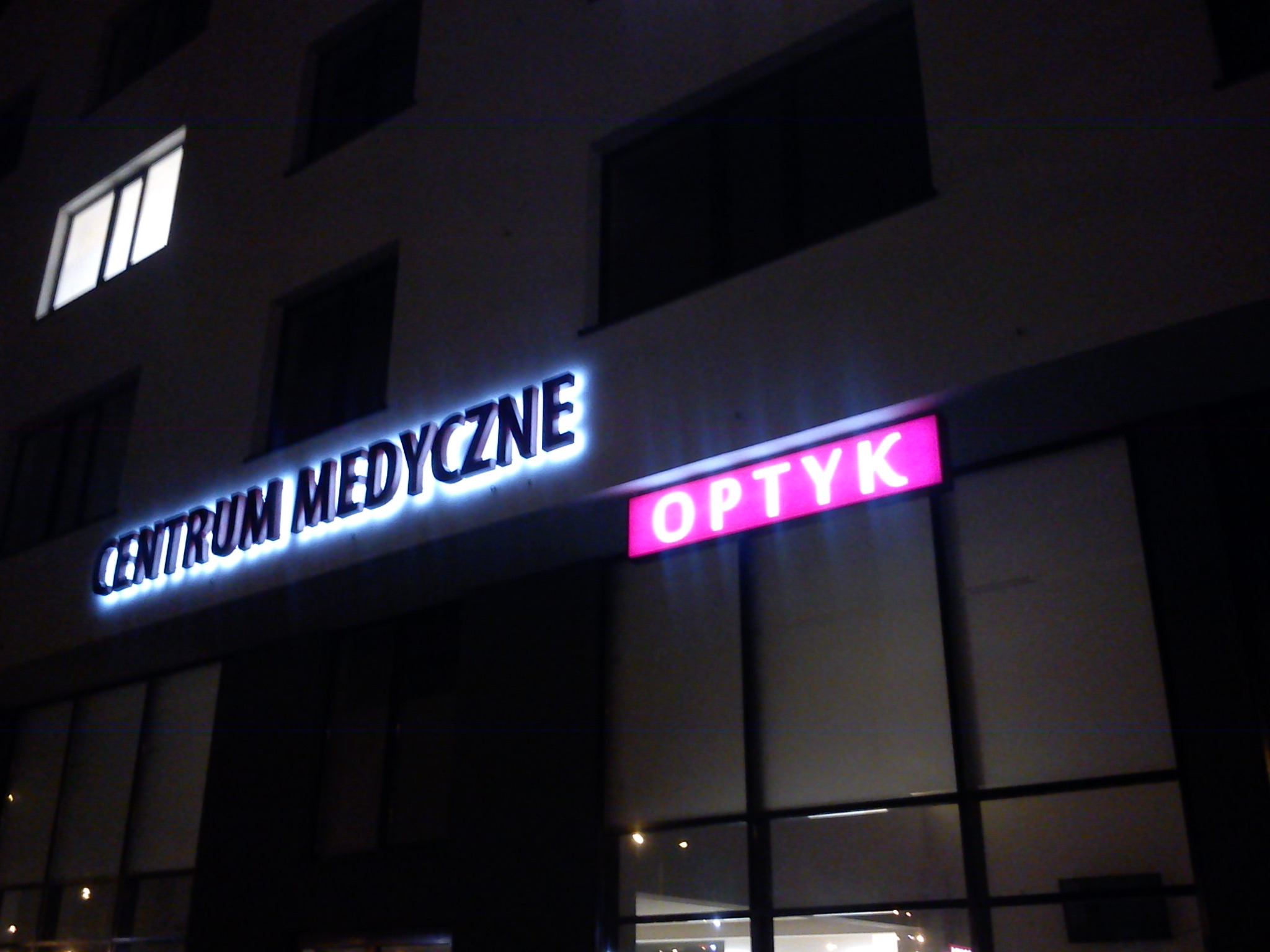 Nocne ujęcie fasady budynku z podświetlanymi szyldami 'Centrum Medyczne' i 'Optyk' nad wejściem.