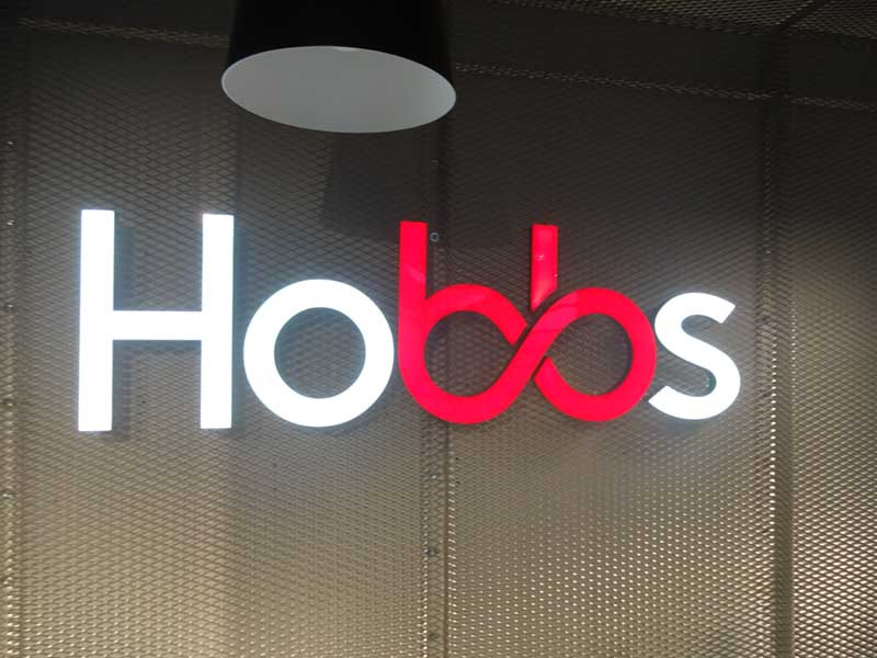 Podświetlane, przestrzenne litery tworzące nazwę 'Hobbs' na metalowej ścianie z fakturą siatki. Litery 'H', 'o' i 's' są białe, a 'bb' czerwone i połączone, tworząc symbol nieskończoności.