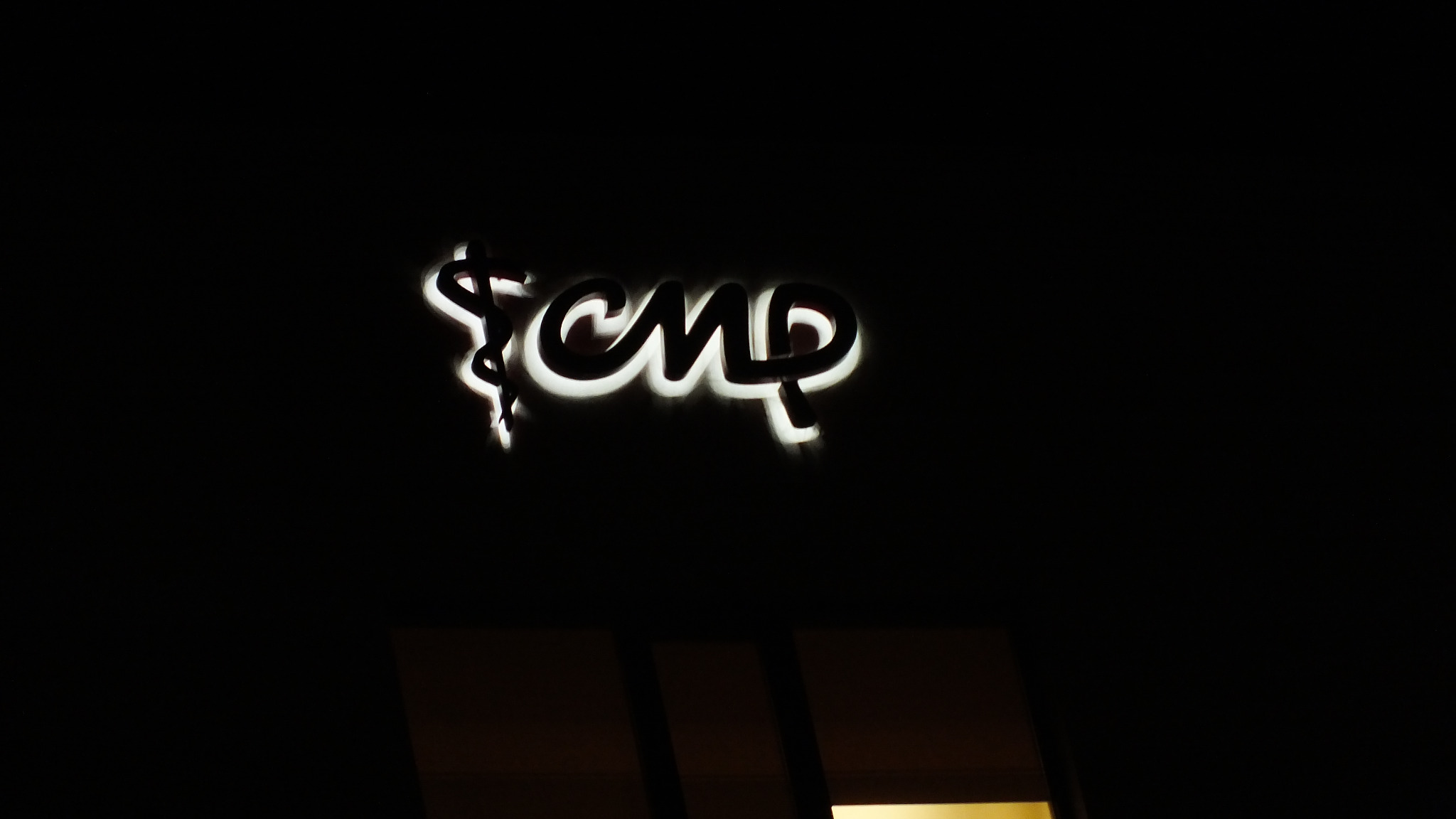 Podświetlany znak z symbolem Eskulapa i napisem CMP na ciemnym tle, widoczne fragmenty okna.